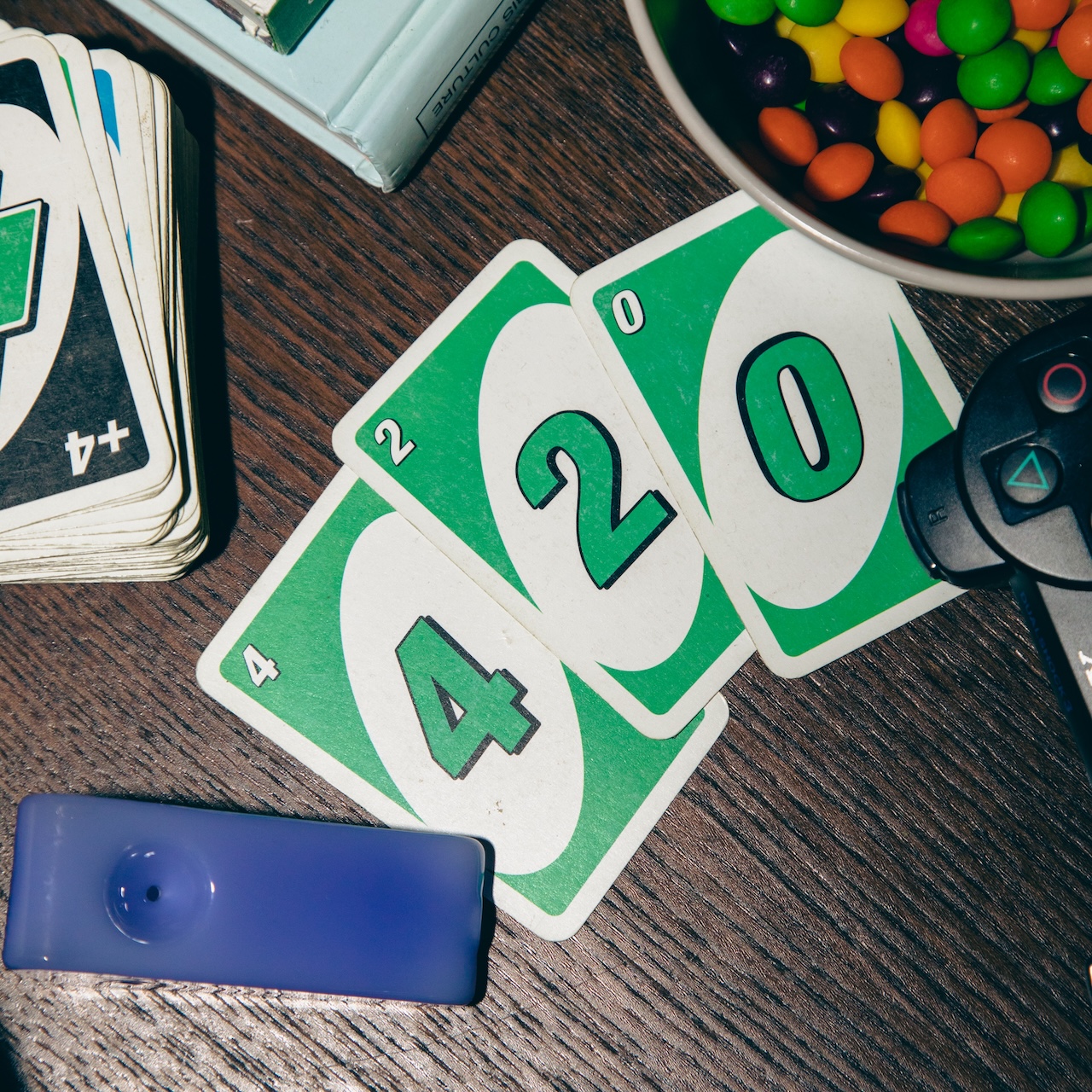 Grüne UNO Karten mit den Zahlen 420 liegen auf einem dunklen Holztisch. Daneben liegt eine blaue Cannabis-Pfeife, bunte Süßigkeiten und ein Gamingcontroller.