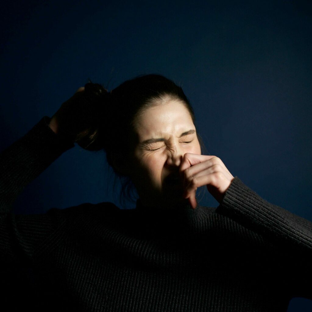 Symbolbild: Frau mit dunklem Pullover niest im Halbdunkel – visuelle Darstellung einer möglichen allergischen Reaktion durch Einatmen von Cannabispartikeln.