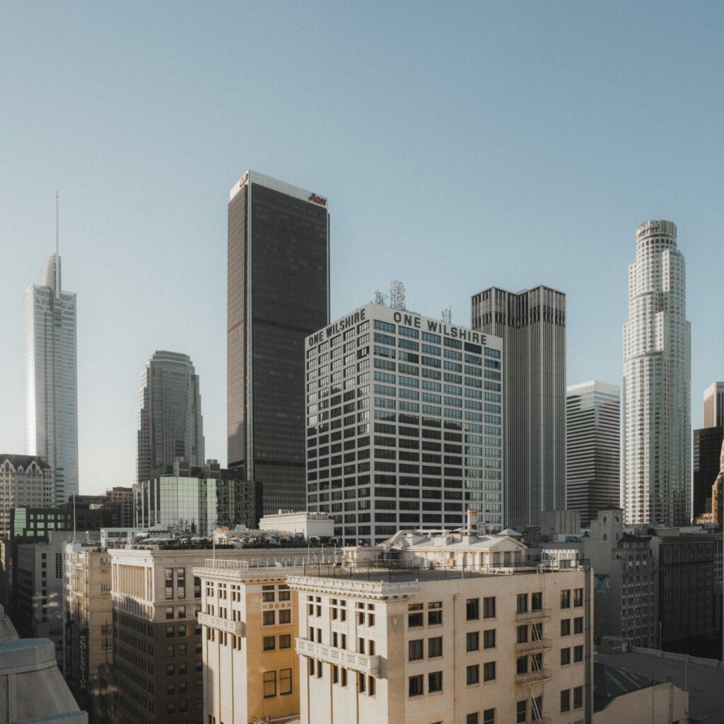Skyline von Downtown Los Angeles bei klarem Tageslicht. Im Vordergrund sind mehrere mittelhohe, beige Gebäude zu sehen, dahinter ragen moderne Wolkenkratzer wie das „One Wilshire“-Gebäude, das US Bank Tower und das Deloitte-Gebäude in den Himmel. Die Architektur spiegelt die urbane Dynamik und wirtschaftliche Bedeutung von L.A. wider – ein Symbolort für Innovation, Popkultur und Cannabis-Trends aus Kalifornien.