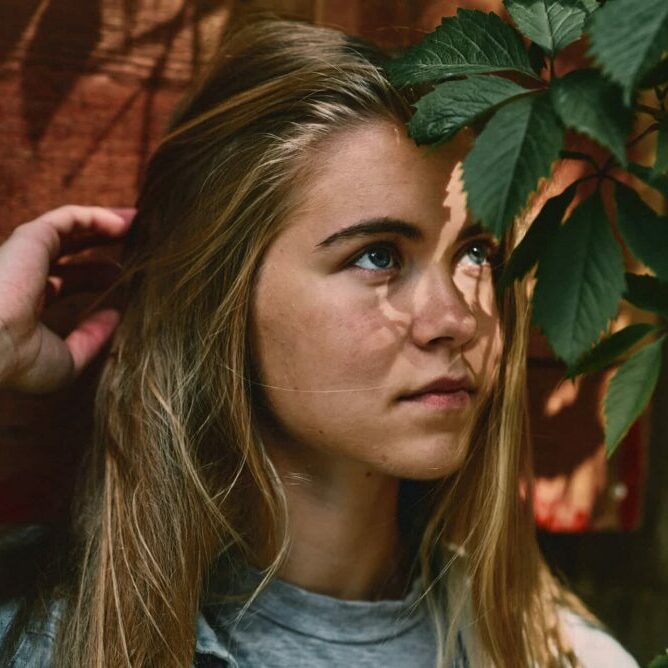 Nachdenkliche junge Frau mit langen blonden Haaren blickt durch grünes Blattwerk – natürliche Licht-Schatten-Komposition als Sinnbild für Selbstreflexion und Achtsamkeit.