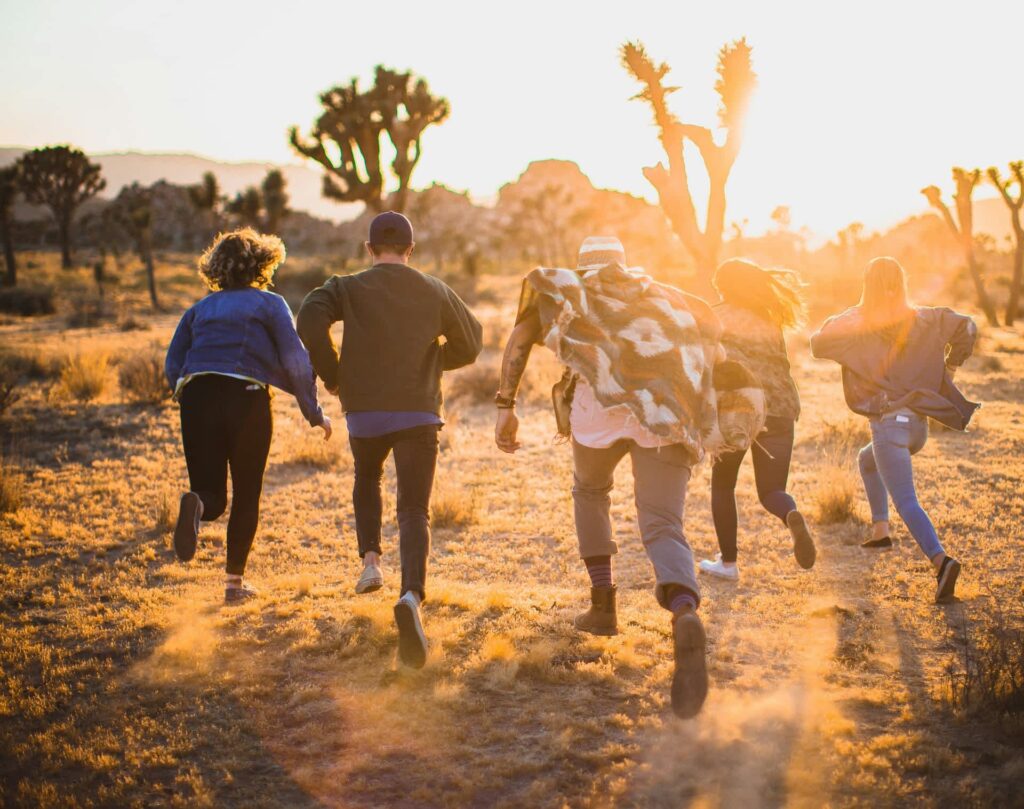 Sechs junge Menschen rennen gemeinsam durch eine sonnendurchflutete Wüstenlandschaft mit kargen Büschen und markanten Joshua Trees. Die Szene ist von warmem, goldenen Licht durchzogen – aufgenommen während des Sonnenuntergangs. Die Gruppe läuft von der Kamera weg, was ein Gefühl von Freiheit, Abenteuerlust und Gemeinschaft vermittelt.